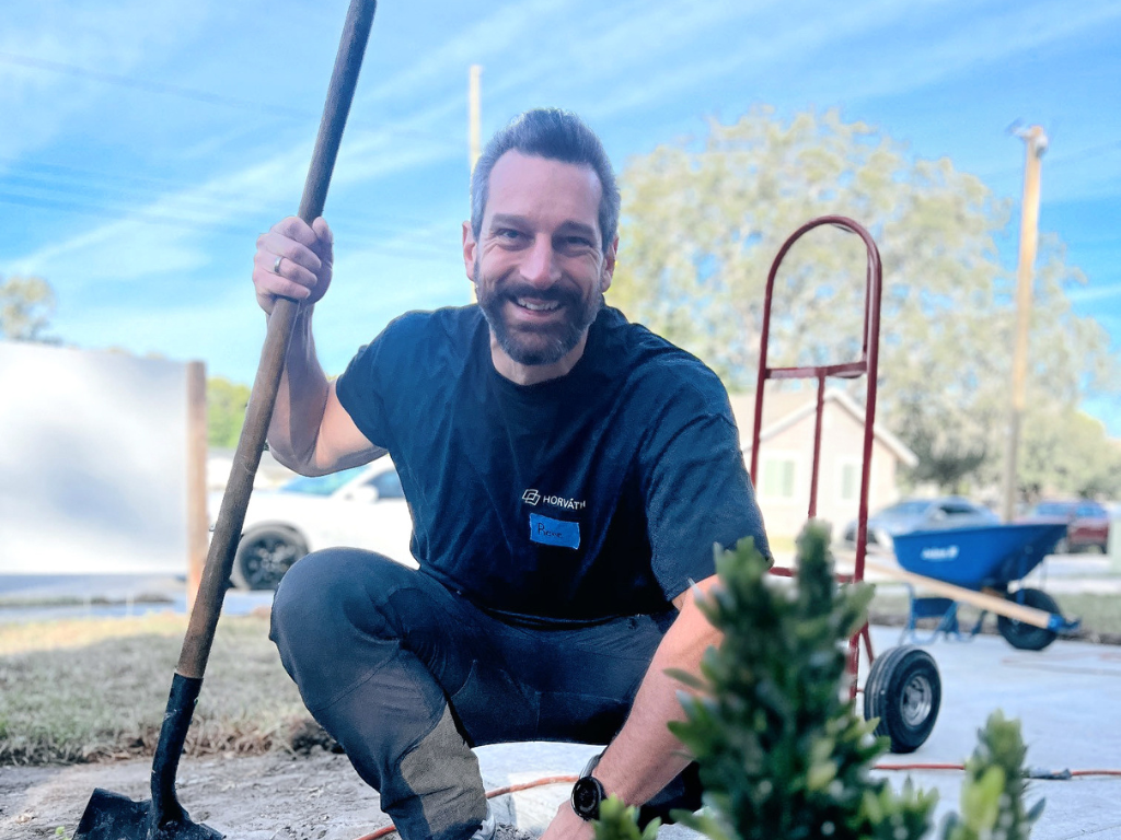 A man in a Horváth shirt kneeling outside with a shovel and garden tools.